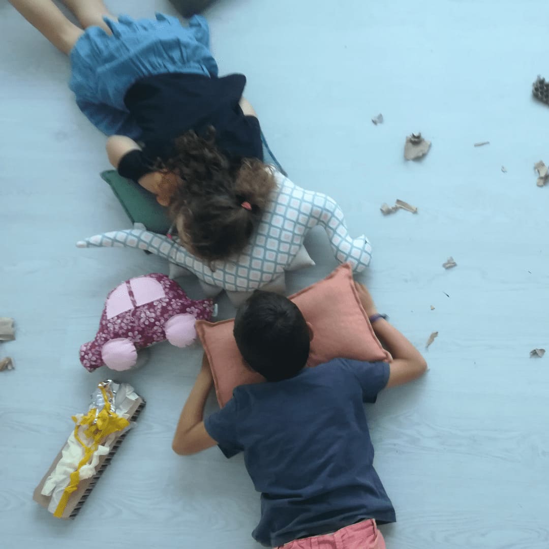Un niño y una niña descansan sobre unos cojines, a su alrededor vemos diferentes materiales usados durante la sesión de arteterapia