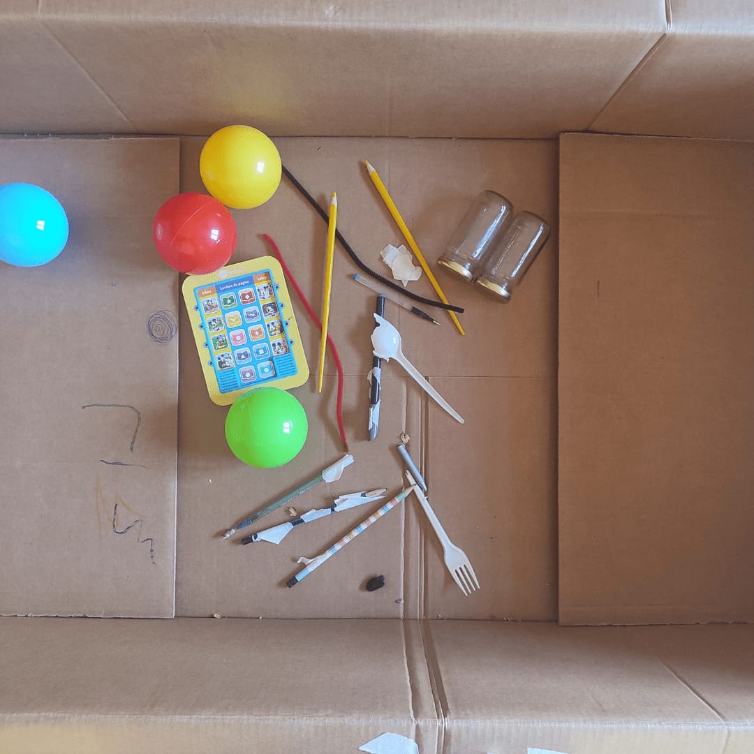 Plano desde arriba de una caja que contiene materiales usados en una sesión de terapia artística integrativa infantil, como lápices, pelotas y tarros.
